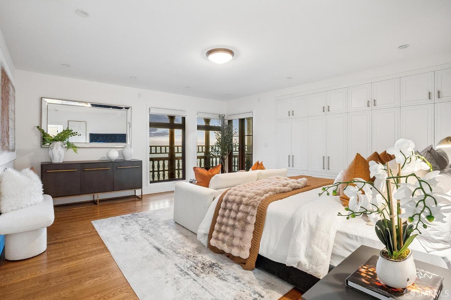 This spacious primary bedroom features a neutral color palette with warm accents, centered around a plush bed dressed in white linens and a textured throw. A large, dark-toned sideboard sits beneath a wide mirror, while built-in white cabinetry provides extensive storage along the wall. The room is brightened by natural light streaming through large windows that offer a view of the outdoors.