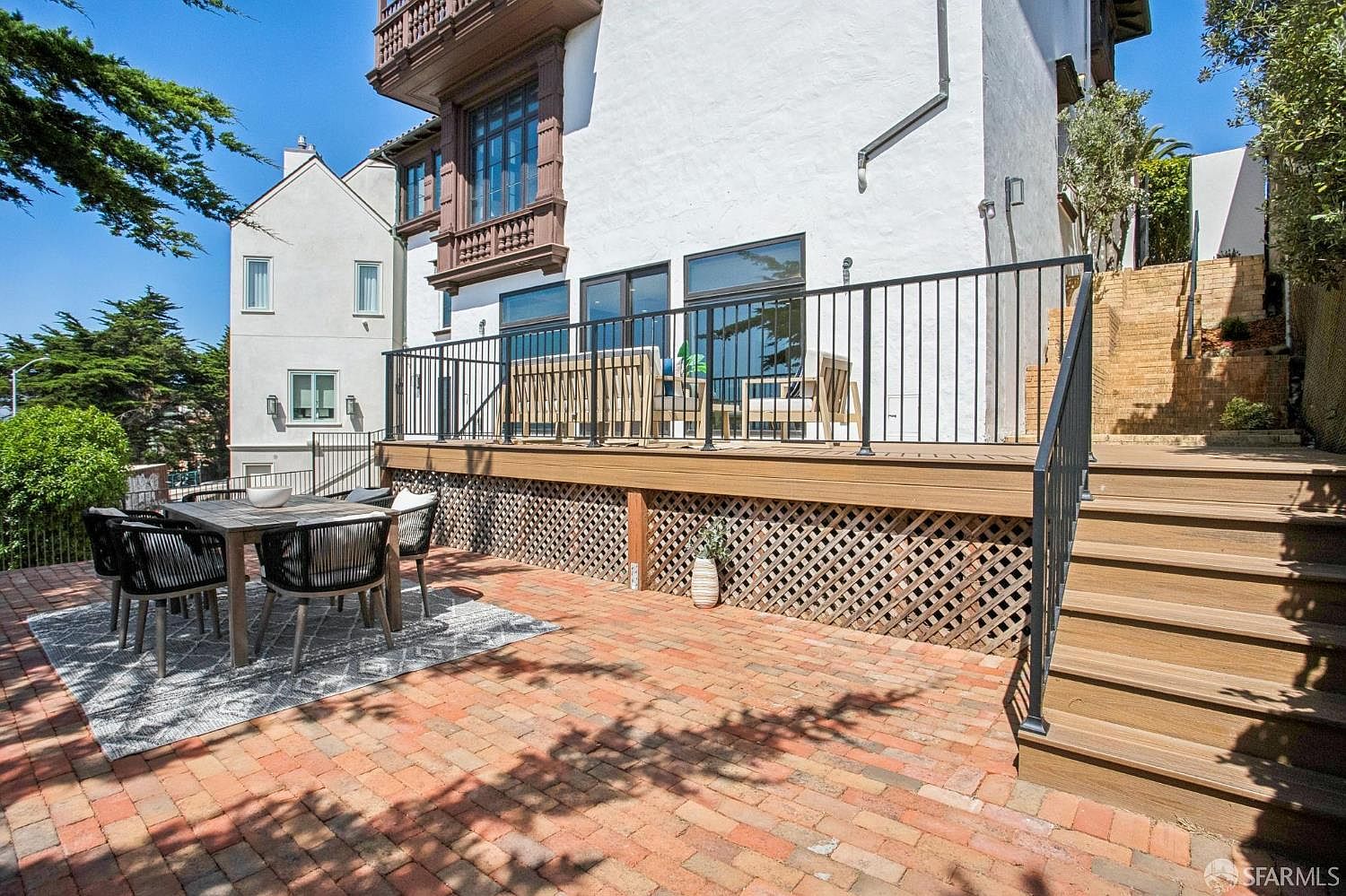This rear exterior view showcases a multi-level outdoor living space featuring a brick-paved patio area with a dining set and a raised wooden deck above. The deck is enclosed by a black metal railing and includes outdoor seating, while a set of wooden stairs leads up to a higher terraced garden level. The white stucco house exterior provides a bright backdrop, complemented by mature trees and a lattice-skirted deck foundation.