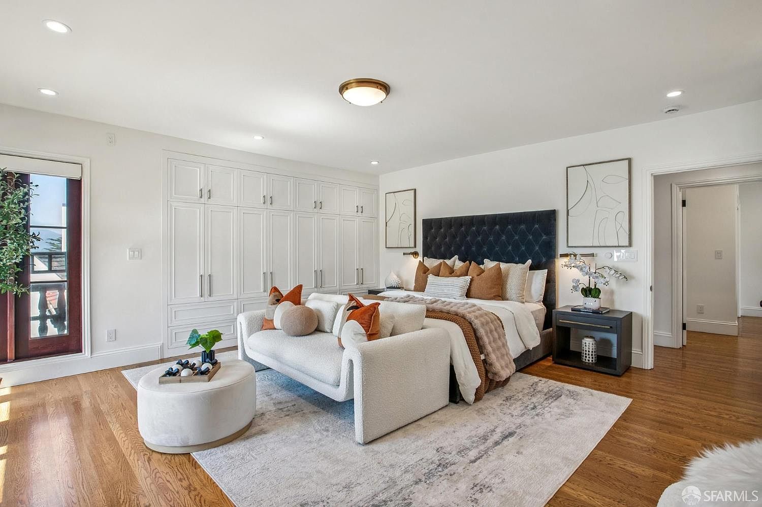 This spacious primary bedroom features a plush, dark-tufted headboard centered against a wall of custom white cabinetry. A light-toned sofa sits at the foot of the bed atop a textured area rug, creating a cozy seating area, while warm wood flooring runs throughout the room. The space is brightened by recessed lighting and a central flush-mount fixture, with a glass door providing access to an outdoor area.
