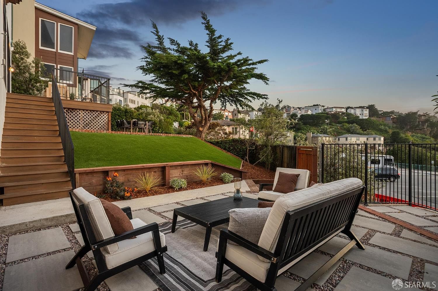 This outdoor living space features a paved patio area furnished with a modern sofa and chairs arranged around a central coffee table. A set of wooden stairs leads up to an elevated deck, while a manicured artificial turf lawn sits atop a retaining wall. The scene is framed by a large, mature tree and a black metal fence, offering a view of the surrounding hillside neighborhood under a twilight sky.