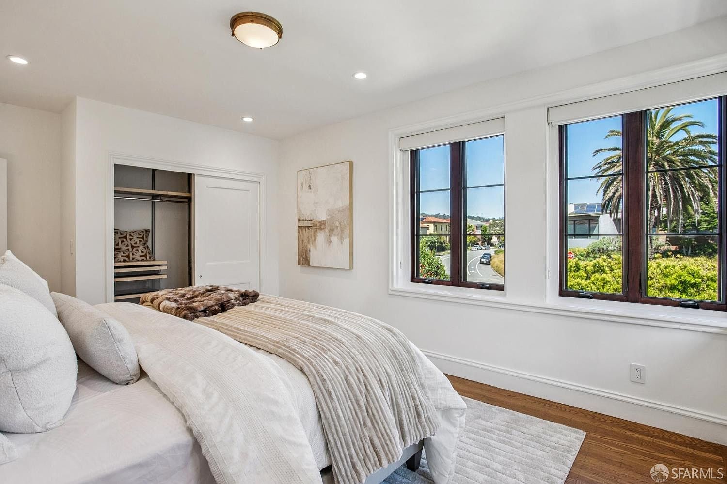 This bright and airy bedroom features a neutral color palette with white walls and warm wood flooring. A plush bed with layered bedding sits adjacent to a walk-in closet and two large windows that provide ample natural light and a view of the neighborhood. The room is accented by a piece of abstract wall art and a simple ceiling light fixture, creating a clean and inviting atmosphere.