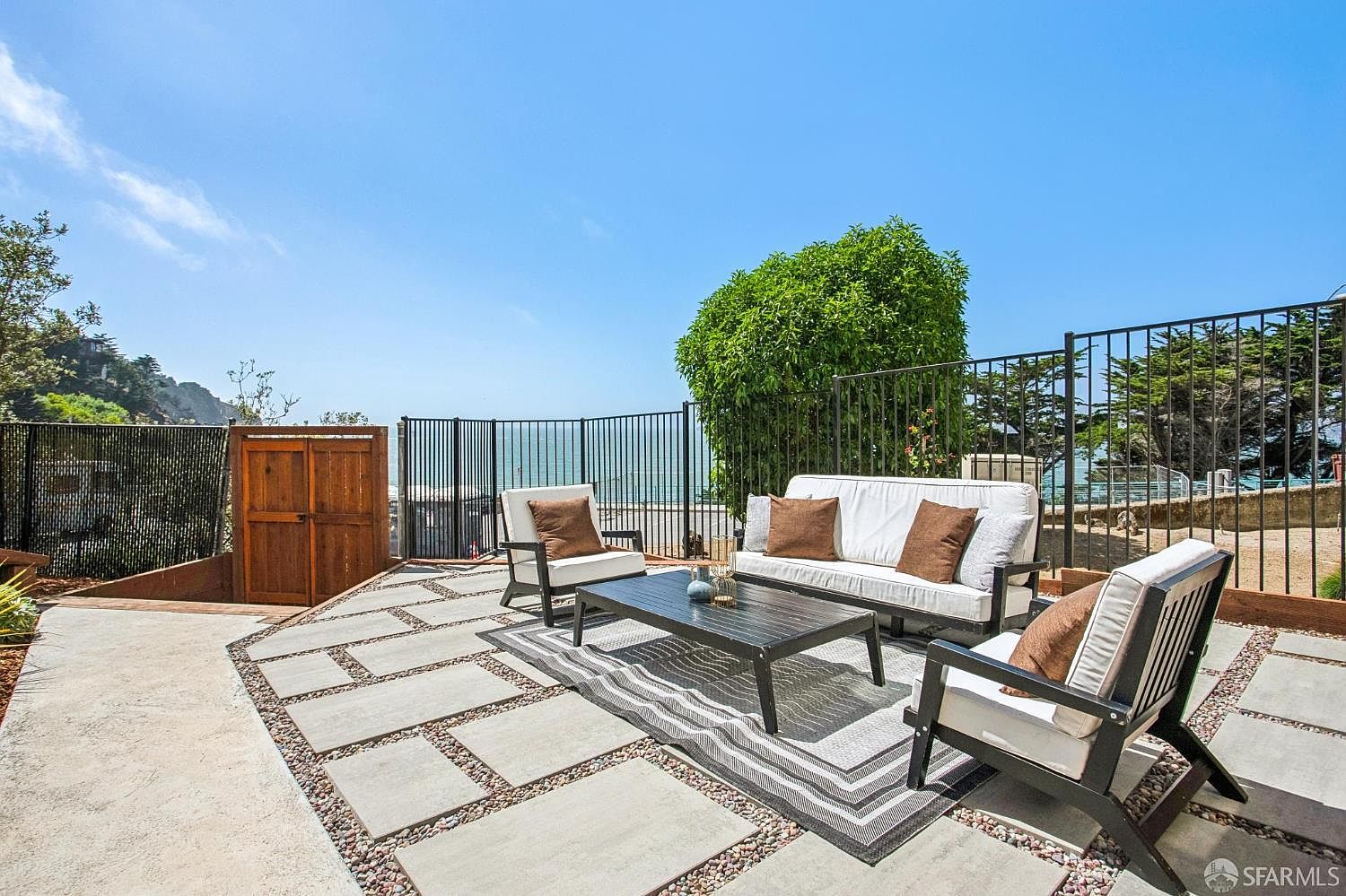 This outdoor patio space features a modern seating arrangement with a white sofa and two armchairs centered around a dark coffee table on a patterned area rug. The patio is constructed with large, light-colored concrete pavers set into a gravel base, providing a clean and contemporary aesthetic. A black metal fence encloses the area, offering an unobstructed view of the ocean and coastline in the distance.