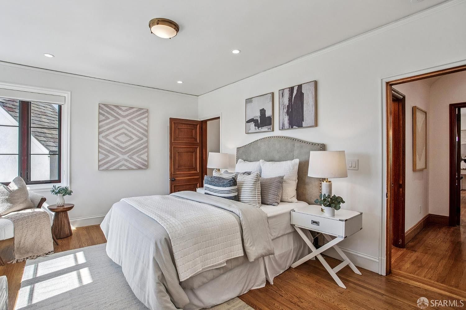 This bright and airy primary bedroom features a neutral color palette with a plush, light-gray upholstered headboard as the focal point. The room is furnished with a comfortable bed dressed in layered linens, a white bedside table with a lamp, and a cozy seating area by the window. Hardwood floors and crisp white walls create a clean, inviting atmosphere, while two abstract art pieces add a touch of modern sophistication.