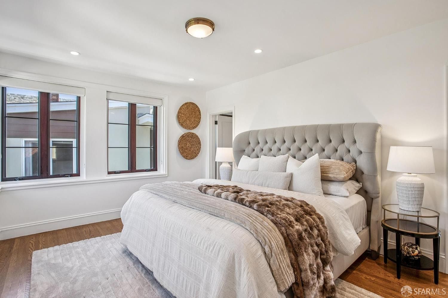 This bright and airy primary bedroom features a large, tufted grey headboard as the focal point, dressed with plush white bedding and a textured brown throw blanket. The room is accented by two circular woven wall decorations and matching bedside tables with white lamps, all set against crisp white walls and warm wood flooring. Large windows with integrated blinds allow for ample natural light, creating a serene and inviting atmosphere.