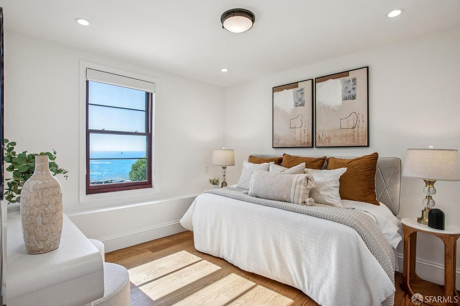 This bright and airy primary bedroom features a plush bed dressed in neutral linens with warm-toned accent pillows. A large window provides a stunning, unobstructed view of the ocean, filling the room with natural light. The space is completed by a side table with a glass-based lamp and abstract wall art, creating a serene and inviting coastal atmosphere.