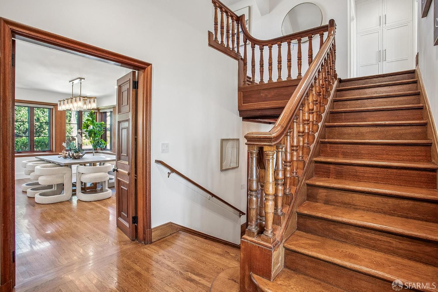 This interior view captures a grand wooden staircase with ornate balusters leading to an upper level, set against a bright, neutral-toned hallway. To the left, an open doorway reveals a dining room featuring a modern chandelier and a table with upholstered chairs. The perspective emphasizes the architectural character of the home, highlighting the contrast between the rich wood tones of the staircase and the clean, airy atmosphere of the foyer.