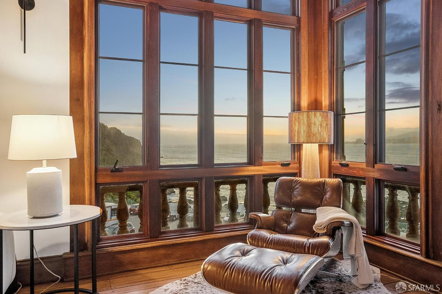 This cozy seating area features a classic leather lounge chair with an ottoman, positioned to take advantage of expansive, multi-pane windows that frame a stunning ocean view. The room is accented by a round side table with a modern lamp and a textured rug, creating a warm and inviting atmosphere. The rich wood trim around the windows adds a traditional touch to this serene space, which is bathed in the soft, golden light of a sunset.