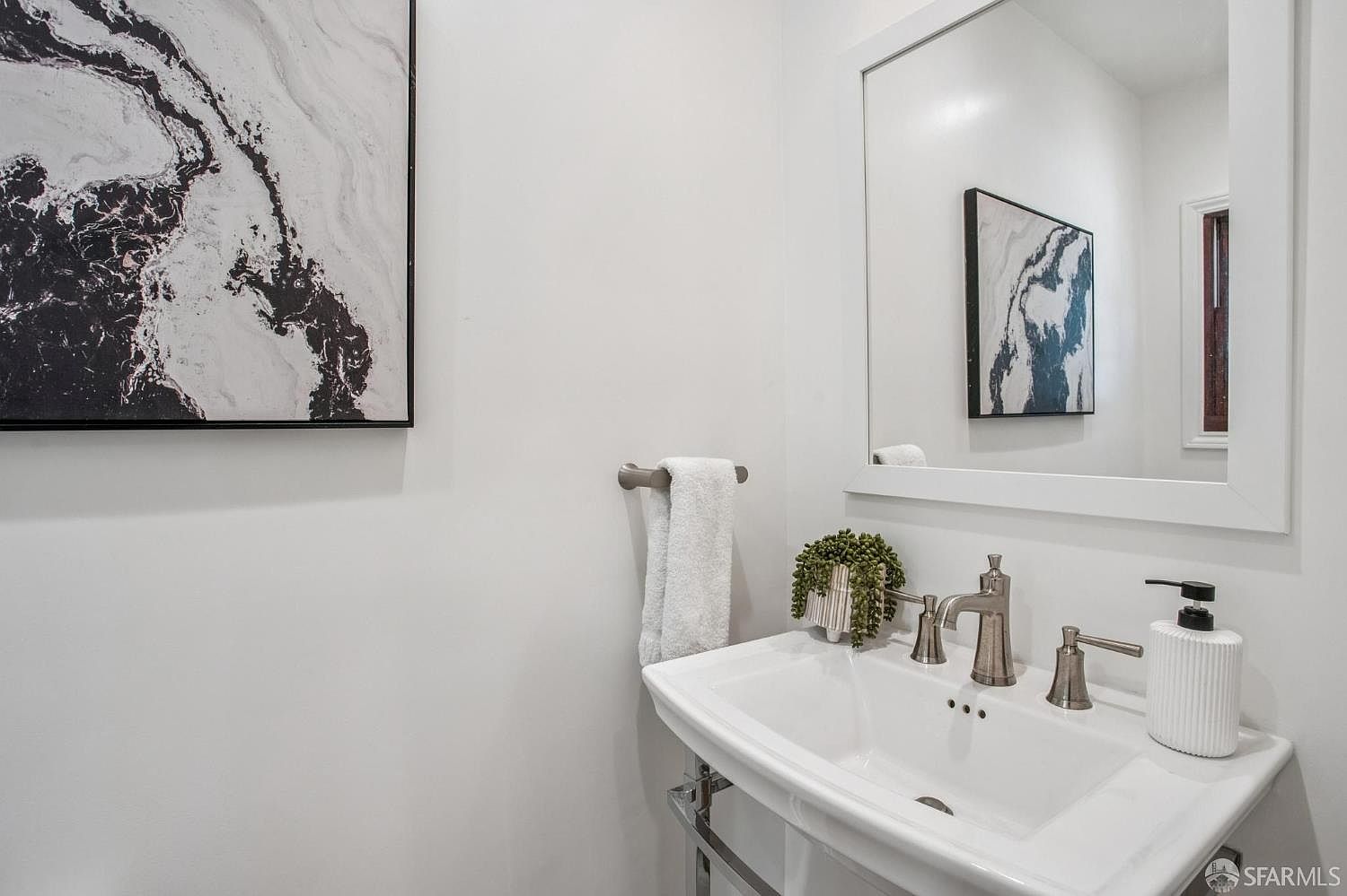 This bright and minimalist powder room features a pedestal sink with a polished metal faucet and a matching towel bar. A large, framed mirror hangs above the sink, reflecting a piece of abstract wall art that is also displayed on the adjacent wall. The space is accented with a small potted plant and a white soap dispenser, creating a clean and modern aesthetic.