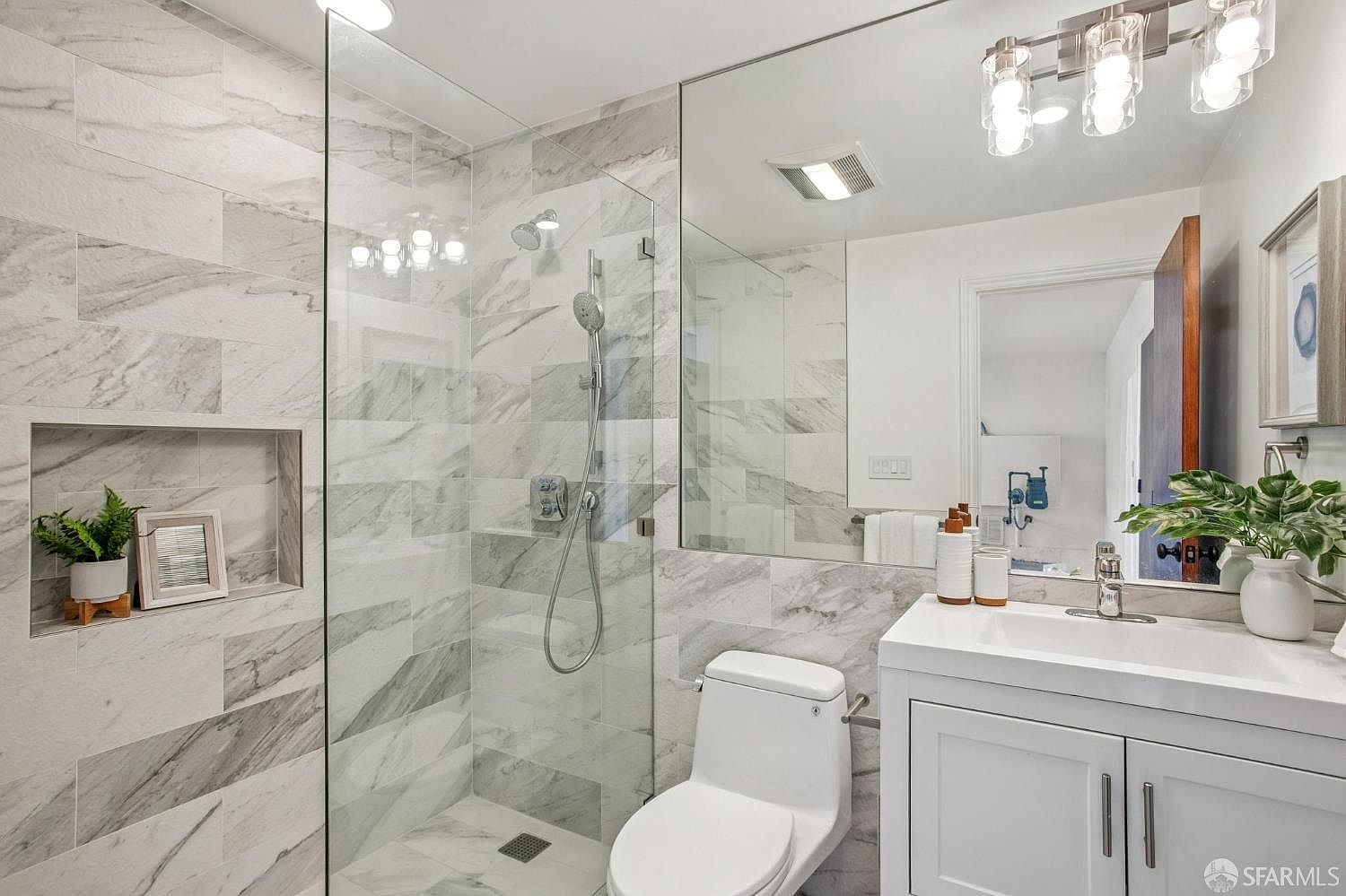 This modern bathroom features a walk-in shower enclosed by a glass panel, showcasing light-toned patterned tile walls and a built-in recessed niche for storage. A white vanity with a clean countertop sits beside a toilet, complemented by a large mirror that enhances the sense of space. The room is illuminated by a contemporary multi-light fixture, creating a bright and polished aesthetic.