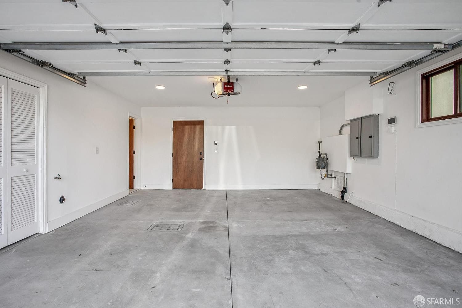 This is a clean, spacious two-car garage featuring white walls, a smooth concrete floor, and an overhead garage door mechanism. The space includes a wooden interior door, a louvered closet door, and a window on the right wall, with utility meters and an electrical panel mounted on the wall. The perspective is a wide-angle shot from the garage entrance, showcasing the functional and well-maintained condition of the area.