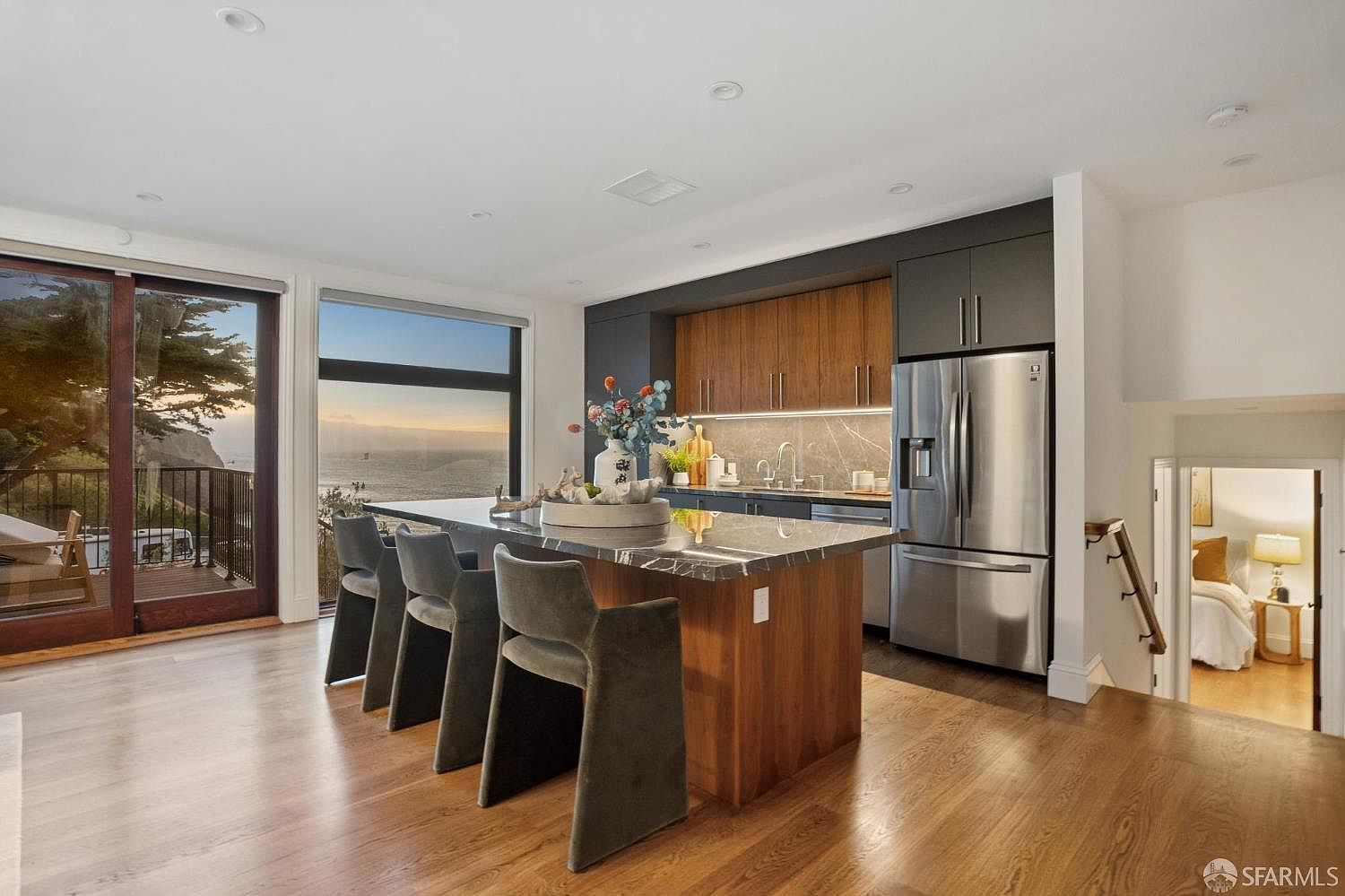 This modern kitchen features a large central island with a stone countertop and seating for three, complemented by warm wood cabinetry and dark charcoal upper cabinets. The space is bathed in natural light from large windows that frame a stunning ocean view, seamlessly connecting the interior to an outdoor deck. A staircase leads down to a secondary living area, creating an open and airy atmosphere with a contemporary, coastal aesthetic.