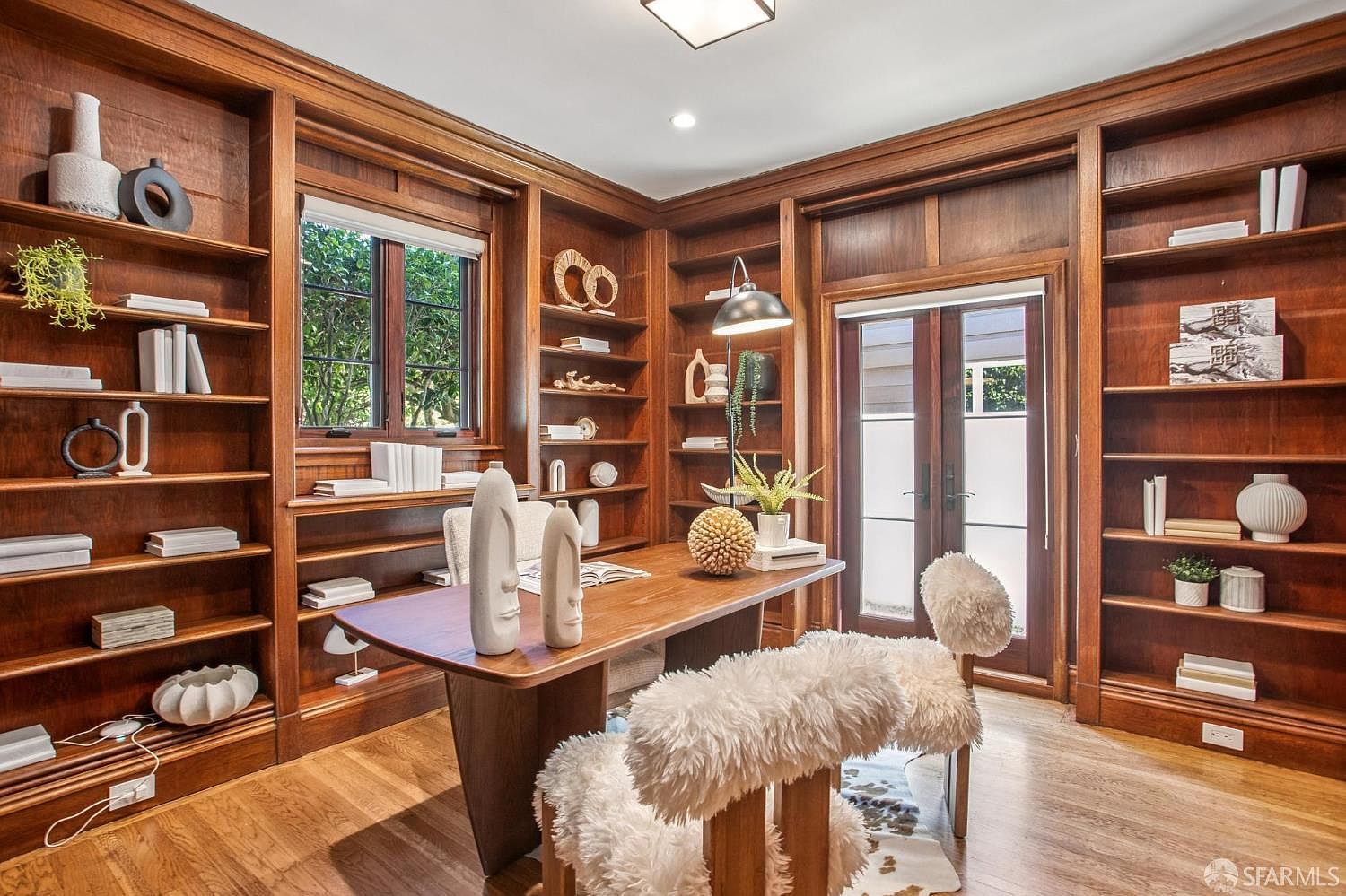 This sophisticated home office features extensive custom wood shelving that wraps around the walls, providing ample storage and display space. A central wooden desk is accented by two unique, textured white chairs, while a large window and glass-paneled doors allow natural light to fill the room. The warm wood tones and thoughtful decor create a professional yet inviting atmosphere suitable for a dedicated workspace.