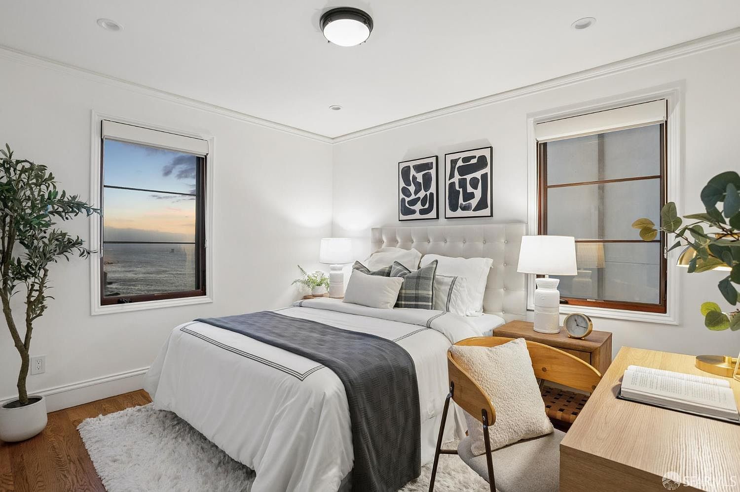 This bright and airy bedroom features a queen-sized bed with a tufted headboard, dressed in crisp white linens and a dark gray throw. The room is framed by two large windows that offer a stunning, unobstructed view of the ocean at sunset. A light wood desk and chair are positioned to the side, creating a functional workspace, while abstract black-and-white artwork adds a modern touch to the minimalist decor.