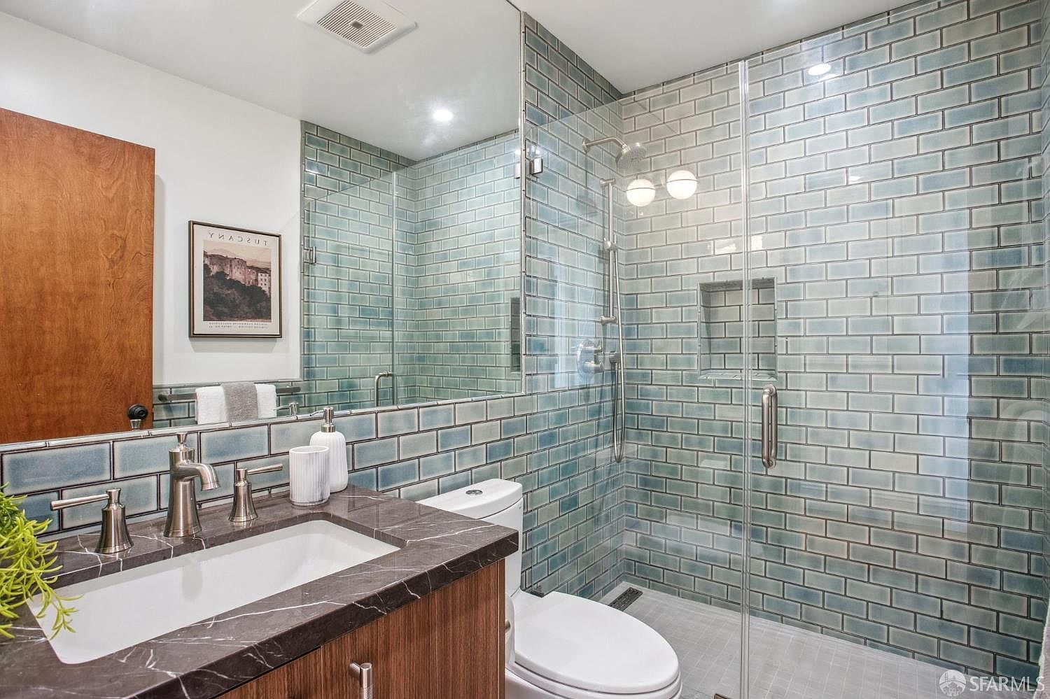 This modern bathroom features a sleek vanity with a dark stone countertop and a glass-enclosed walk-in shower. The walls are adorned with glossy, blue-toned subway tiles that extend into the shower area, creating a cohesive and refreshing aesthetic. A large mirror above the vanity enhances the sense of space, while the minimalist fixtures and clean lines contribute to a contemporary, spa-like atmosphere.
