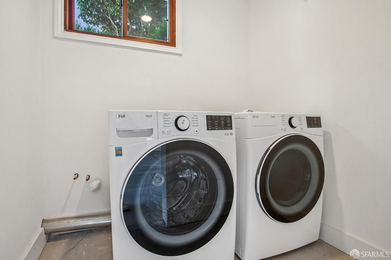 This functional laundry room features a side-by-side LG ThinQ washer and dryer set positioned against a plain white wall. A window above the appliances allows natural light into the space, which has simple, utilitarian finishes. The room provides a clean and straightforward layout suitable for household chores.