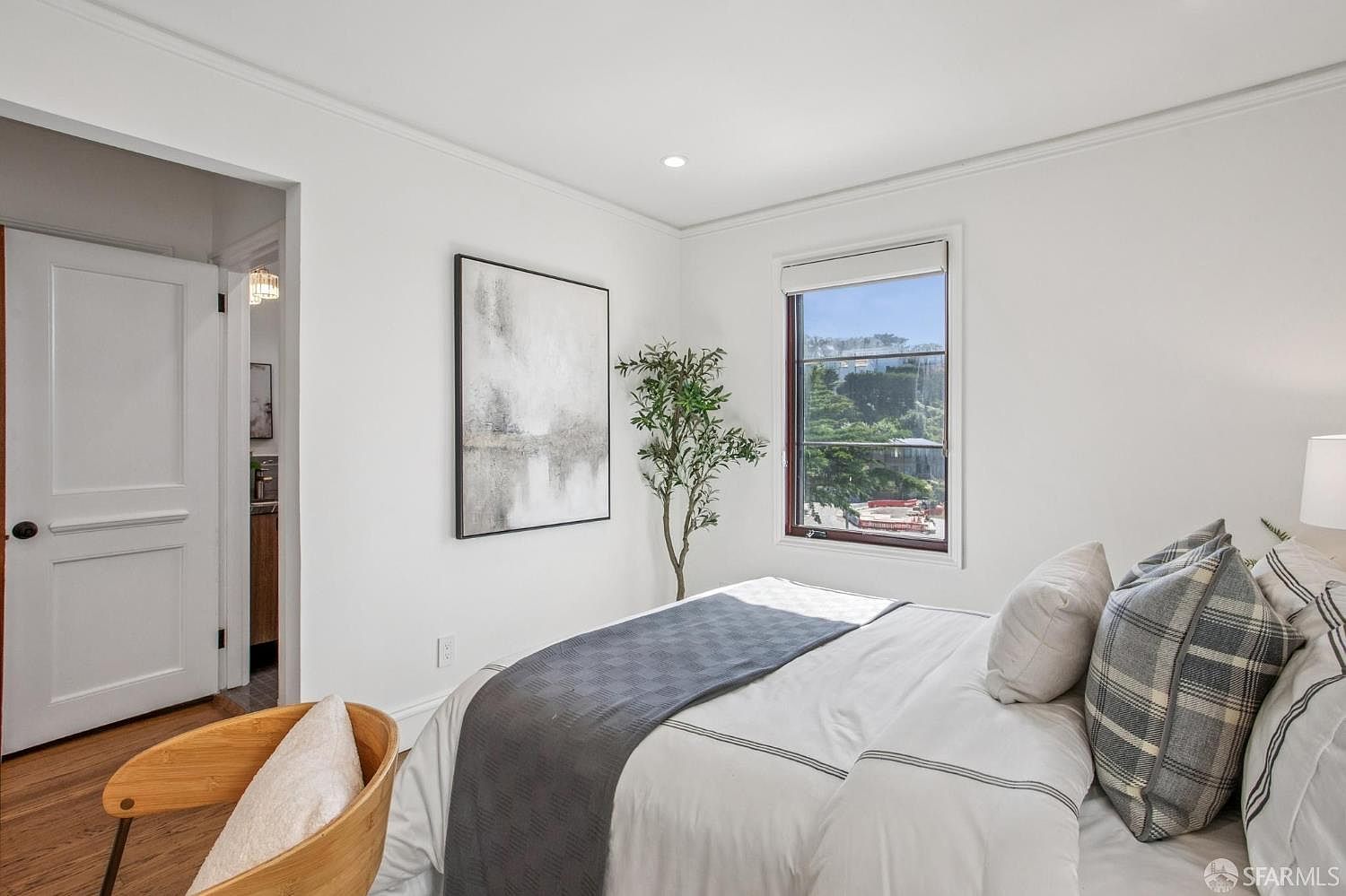 This bright and airy bedroom features crisp white walls, hardwood flooring, and a window that provides a view of the surrounding neighborhood. The room is furnished with a bed dressed in neutral linens with a dark grey runner and plaid accent pillows, complemented by a light wood chair in the foreground and a potted plant in the corner. A doorway leads to an adjacent bathroom, creating a cohesive and inviting living space.