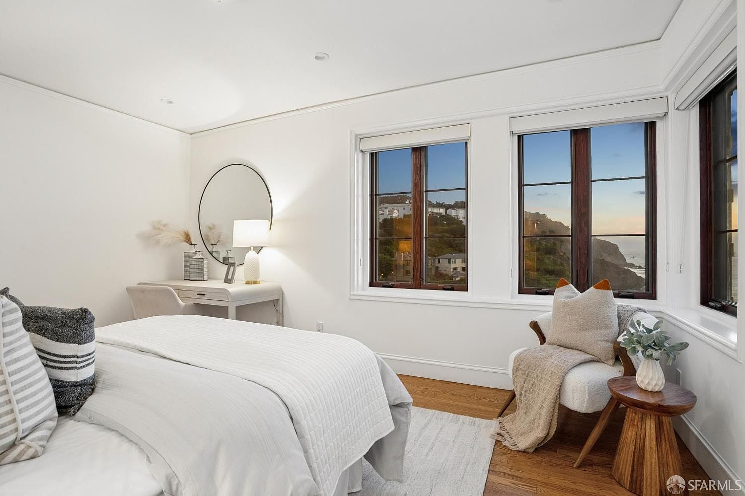 This bright and airy bedroom features a neutral color palette with white walls and light wood flooring. The room is furnished with a comfortable bed, a small vanity area with a circular mirror, and a cozy reading nook positioned by large windows that offer a scenic view of the coastline. The space feels serene and inviting, emphasizing natural light and a clean, modern aesthetic.