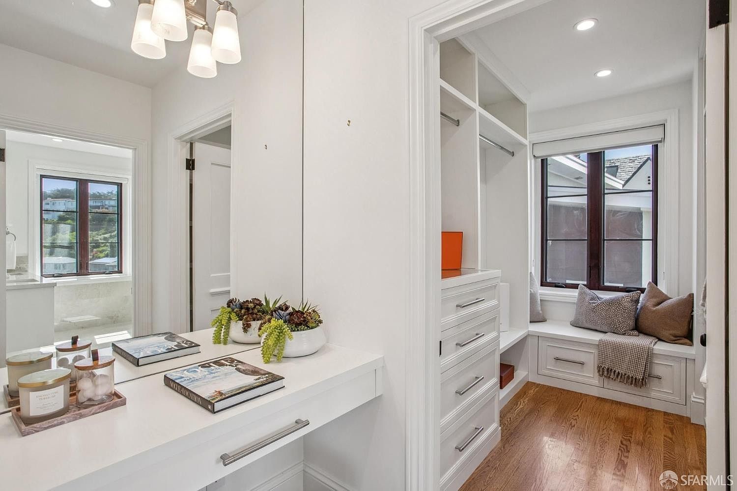 This bright walk-in closet features a built-in vanity area with a large mirror, white cabinetry, and a dedicated seating nook by the window. The space is finished with light wood flooring and custom shelving, creating a clean and organized aesthetic. The perspective looks from the vanity area into the closet nook, highlighting the functional layout and natural light.