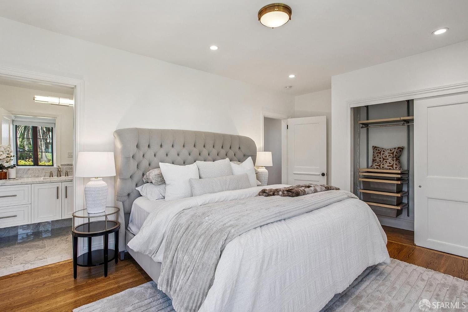 This primary bedroom features a large, tufted grey upholstered headboard as the focal point, complemented by a neutral color palette and soft textures. The room includes a bedside table with a lamp, a view into an adjacent bathroom, and an open-concept closet area with built-in shelving. The space is bright and airy, with hardwood flooring and a plush area rug creating a comfortable and inviting atmosphere.