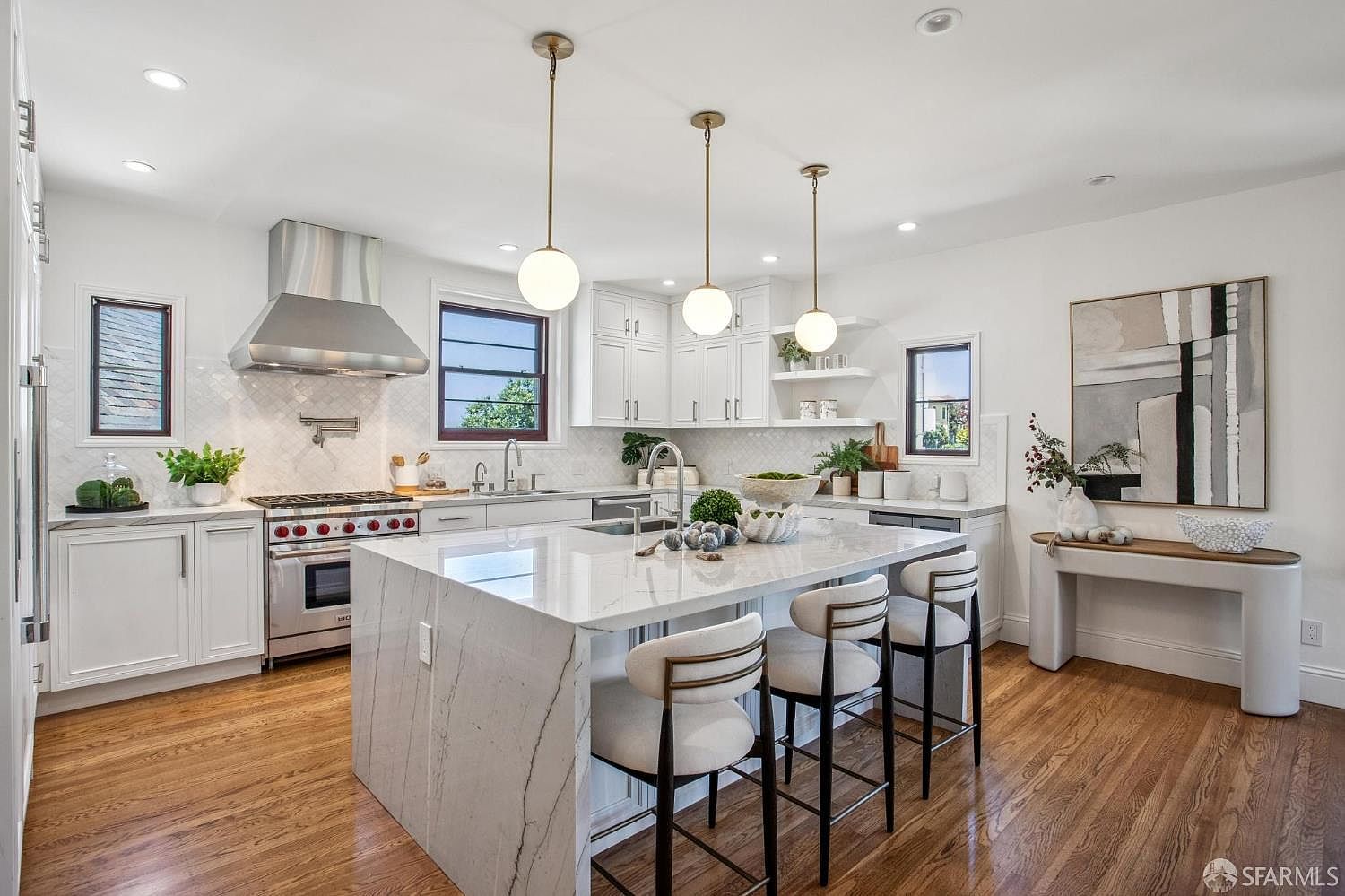 This bright and airy kitchen features white cabinetry, a large central island with a stone waterfall edge, and warm wood flooring. The space is illuminated by three modern pendant lights hanging over the island, complemented by stainless steel appliances and a professional-style range hood. A console table with abstract artwork adds a decorative touch to the side wall, creating a sophisticated and inviting atmosphere.