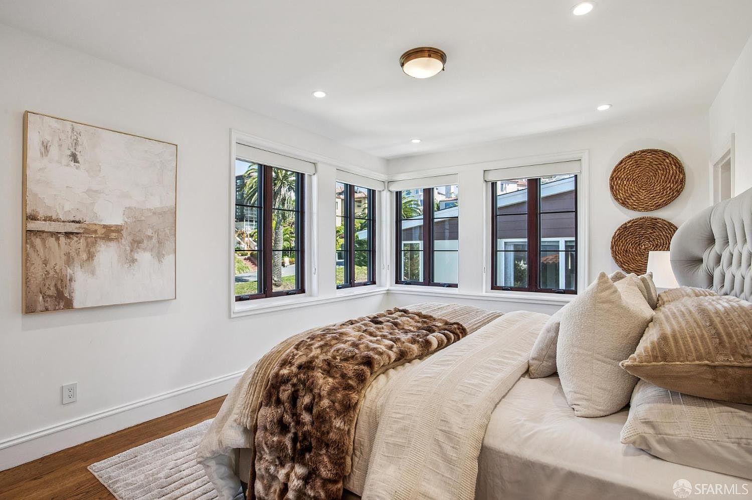 This bright and airy primary bedroom features a plush bed with a tufted headboard, layered with neutral-toned bedding and a textured faux-fur throw. Large windows wrap around the corner of the room, allowing for ample natural light and views of the surrounding greenery. The space is accented by minimalist wall art and circular woven wall decor, creating a serene and sophisticated atmosphere.