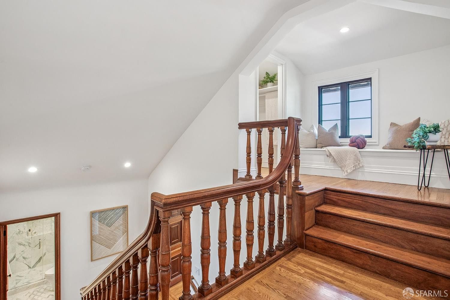 This interior view captures a staircase landing featuring a rich-toned wooden banister and matching hardwood flooring. A cozy window seat is nestled under a window, providing a bright nook that overlooks the landing, while a glimpse of a bathroom is visible to the left. The space is characterized by clean white walls and recessed lighting, creating a bright and inviting transition area.