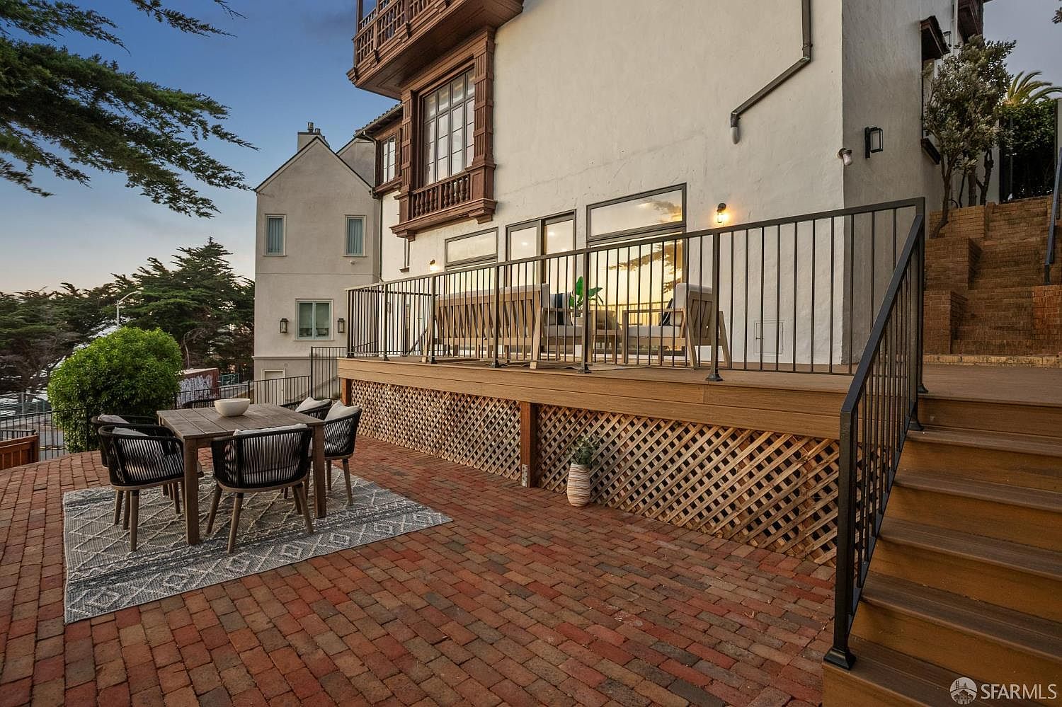This outdoor living space features a multi-level design with a brick-paved patio area and an elevated wooden deck. The deck is enclosed by a black metal railing and includes comfortable outdoor seating, while the patio below is furnished with a dining set atop a patterned rug. The scene is captured from a low angle, highlighting the transition between the hardscape patio and the raised deck against the white stucco exterior of the home.
