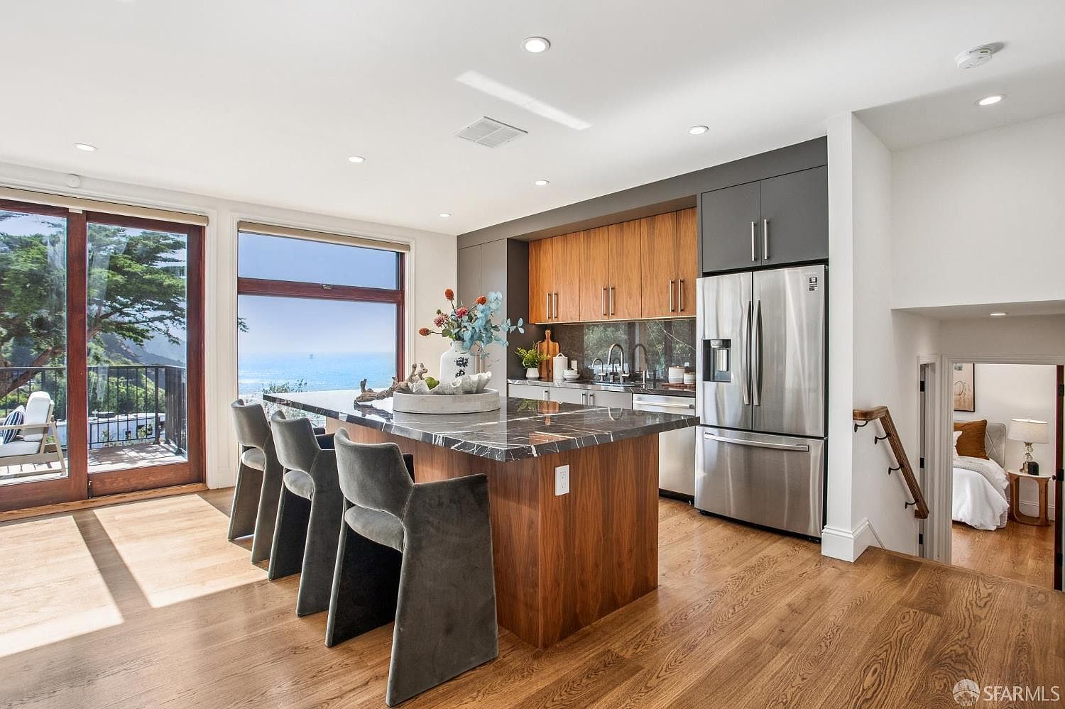 This modern kitchen features a large central island with a dark stone countertop and wood-toned base, paired with three grey upholstered bar stools. The space is highlighted by warm wood cabinetry, stainless steel appliances, and large windows that provide a panoramic view of the ocean. A staircase leads down to a bedroom, creating an open and airy atmosphere that blends contemporary design with coastal scenery.