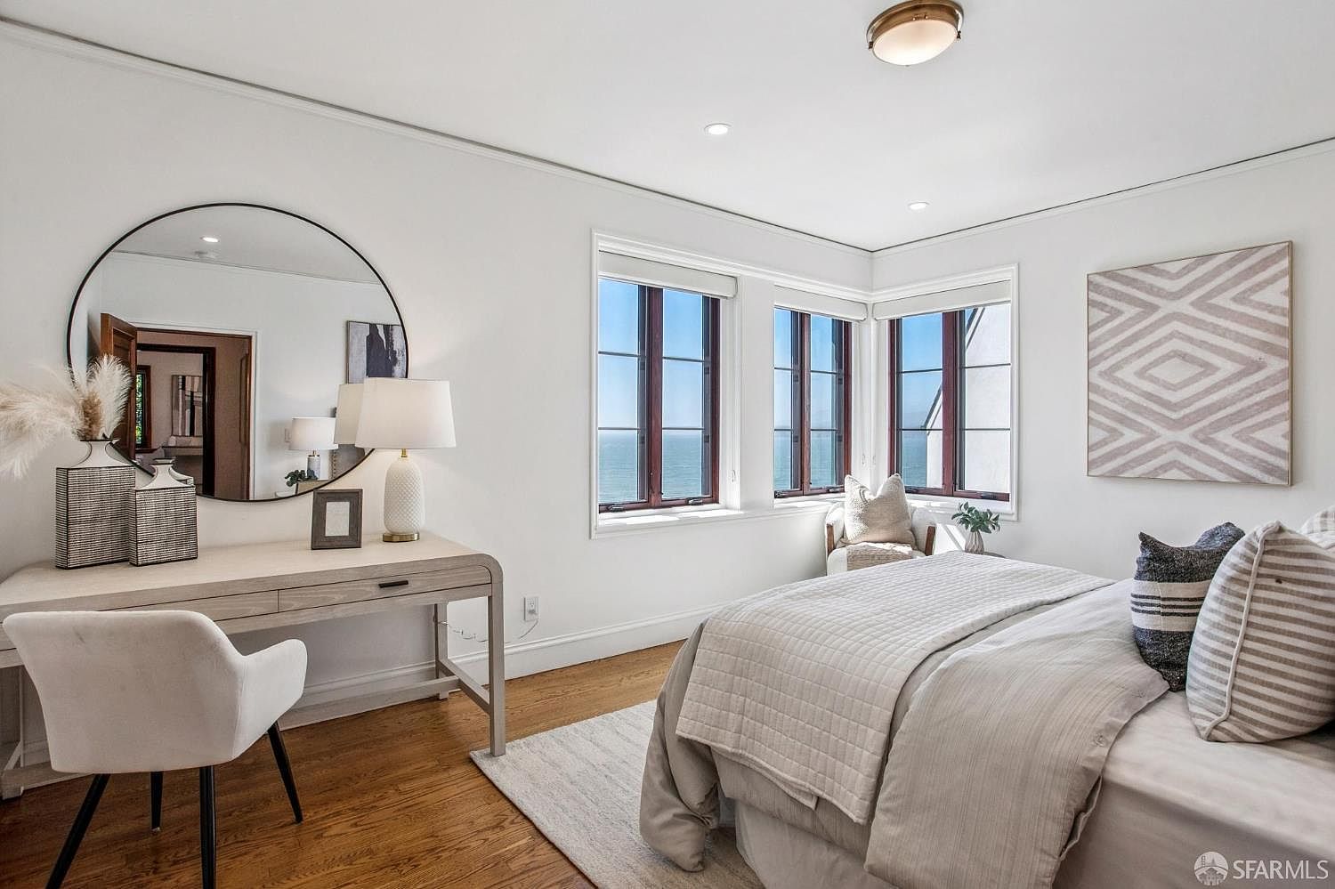 This bright and airy bedroom features a neutral color palette, hardwood flooring, and a large bed dressed in soft linens. A vanity desk with a large circular mirror sits to the left, while a corner window arrangement provides expansive ocean views and natural light. The room is accented by a geometric wall art piece and a cozy seating area by the window, creating a serene and inviting atmosphere.
