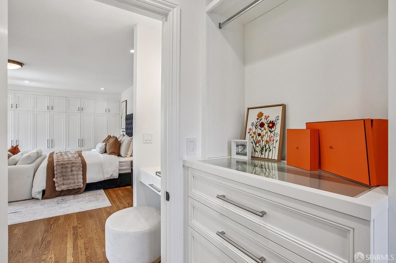 This view captures a walk-in closet area featuring white custom cabinetry with sleek metal hardware and a glass-topped vanity surface. Through the doorway, a bright primary bedroom is visible, showcasing a large bed with neutral-toned bedding and an extensive wall of built-in storage. The perspective creates a seamless transition between the organized closet space and the serene, well-lit bedroom beyond.