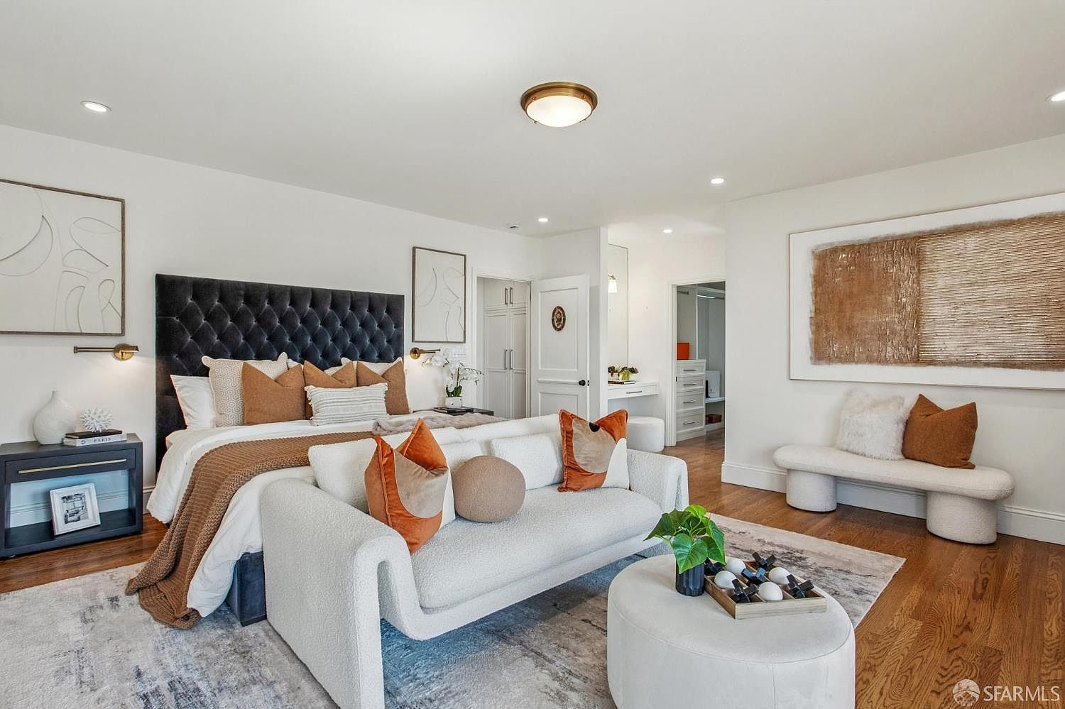 This spacious primary bedroom features a sophisticated design with a tufted dark velvet headboard and a neutral color palette accented by warm terracotta tones. The room includes a cozy seating area with a curved sofa and a matching ottoman, complemented by a textured area rug and a minimalist bench against the wall. Large, abstract artwork and warm lighting fixtures create an inviting and luxurious atmosphere, while the open layout leads into a bright, connected dressing area.