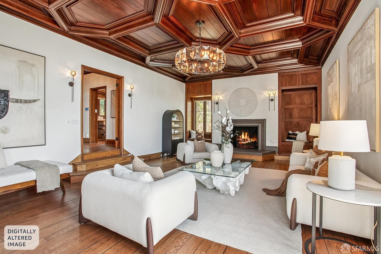 This sophisticated living room features a striking coffered wood ceiling and a central fireplace with a stone surround. The space is furnished with plush, light-toned seating arranged around a unique, organic-shaped coffee table on a neutral area rug. Warm wood flooring and wall sconces create an inviting atmosphere, while a doorway leads to an adjacent room, emphasizing the home's open and elegant flow.