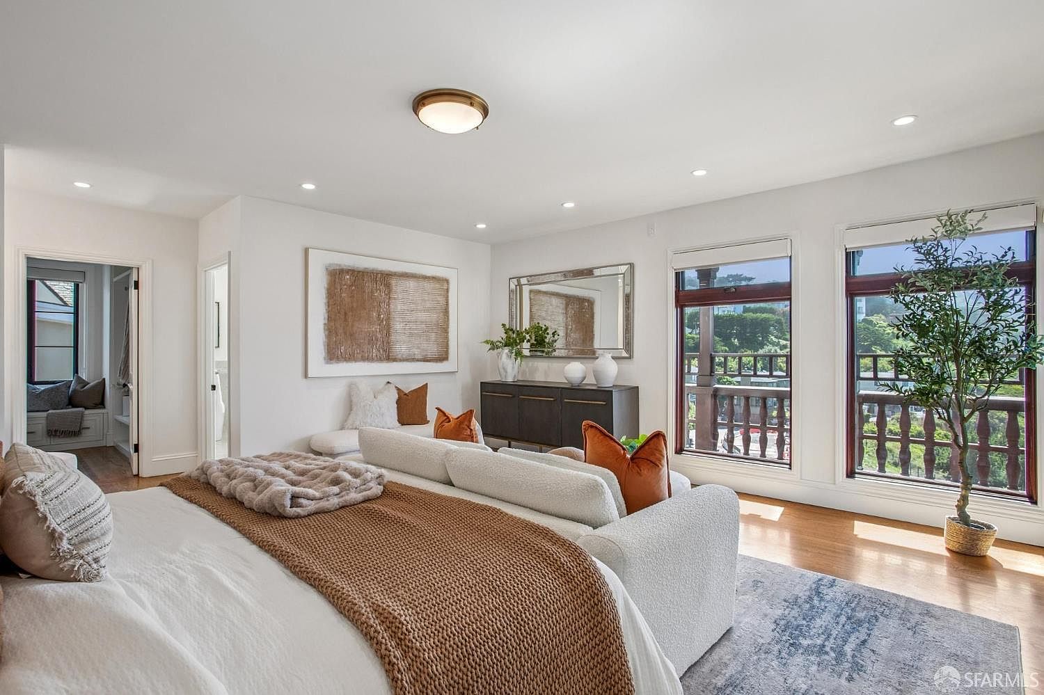 This spacious primary bedroom features a neutral color palette with warm accents, centered around a large bed with a textured brown throw blanket. A sofa is positioned at the foot of the bed, facing a dark wood console table topped with a mirror and decorative vases. Large windows provide ample natural light and views of the surrounding greenery, while a potted tree adds a touch of nature to the corner.