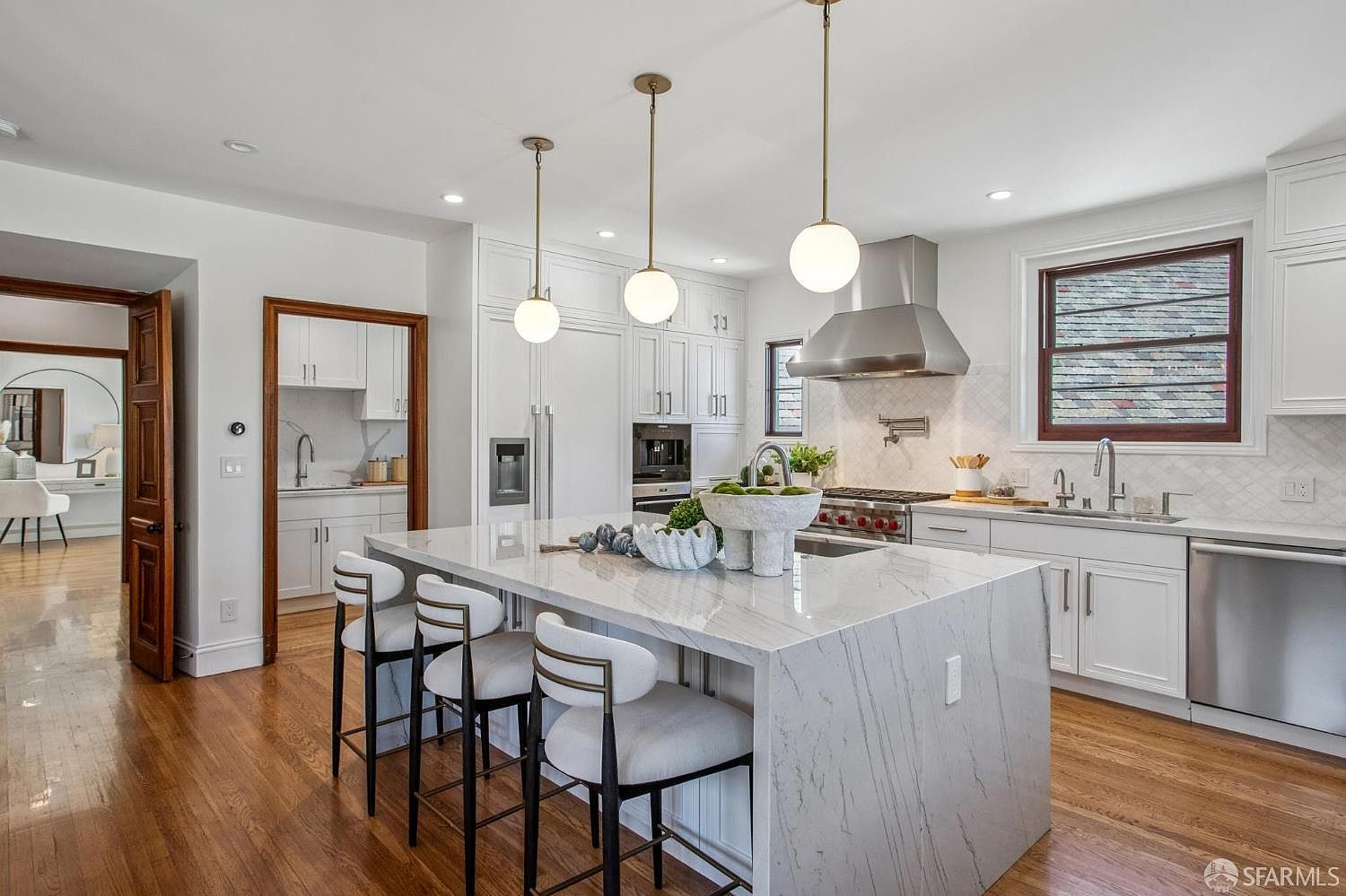This bright, modern kitchen features a large central island with a stone countertop and seating for three, illuminated by three spherical pendant lights. The space is equipped with white cabinetry, stainless steel appliances, and a professional-style range with a metal hood. A doorway leads to a secondary prep area, while warm wood flooring and a window over the sink add a welcoming, functional touch to the design.