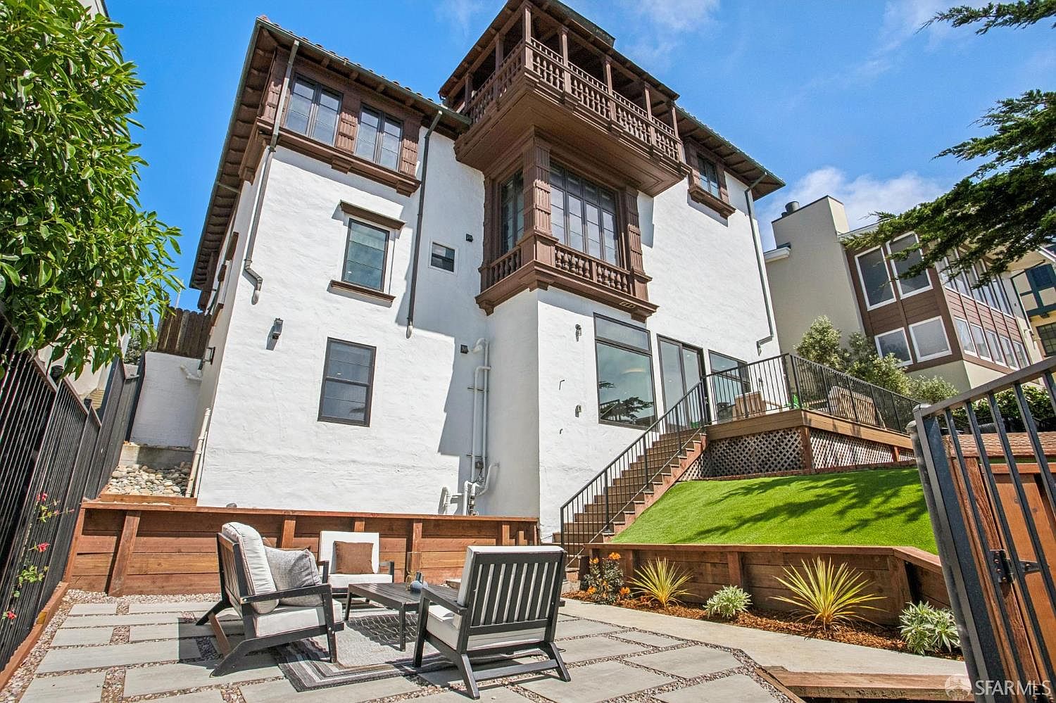 This rear view of a multi-story residence showcases a white stucco facade accented by dark wood trim and a prominent, ornate wooden balcony. The property features a tiered backyard with a stone-paved patio area, a raised wooden deck with black metal railings, and a manicured lawn. The perspective is from the ground level, looking up at the architectural details and the transition between the outdoor living space and the home's rear elevation.