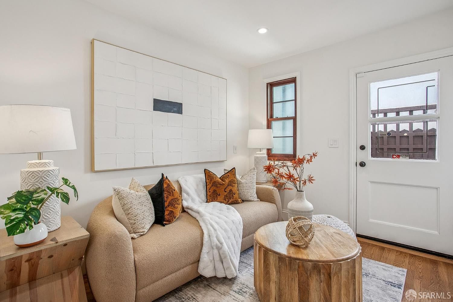 This cozy living room features a neutral-toned curved sofa adorned with patterned and solid throw pillows, draped with a soft white throw blanket. A round light wood coffee table sits on a textured area rug, while a large, minimalist textured wall art piece serves as a focal point above the sofa. The space is illuminated by two matching table lamps and includes a small side table with a potted plant, creating a warm and inviting atmosphere.