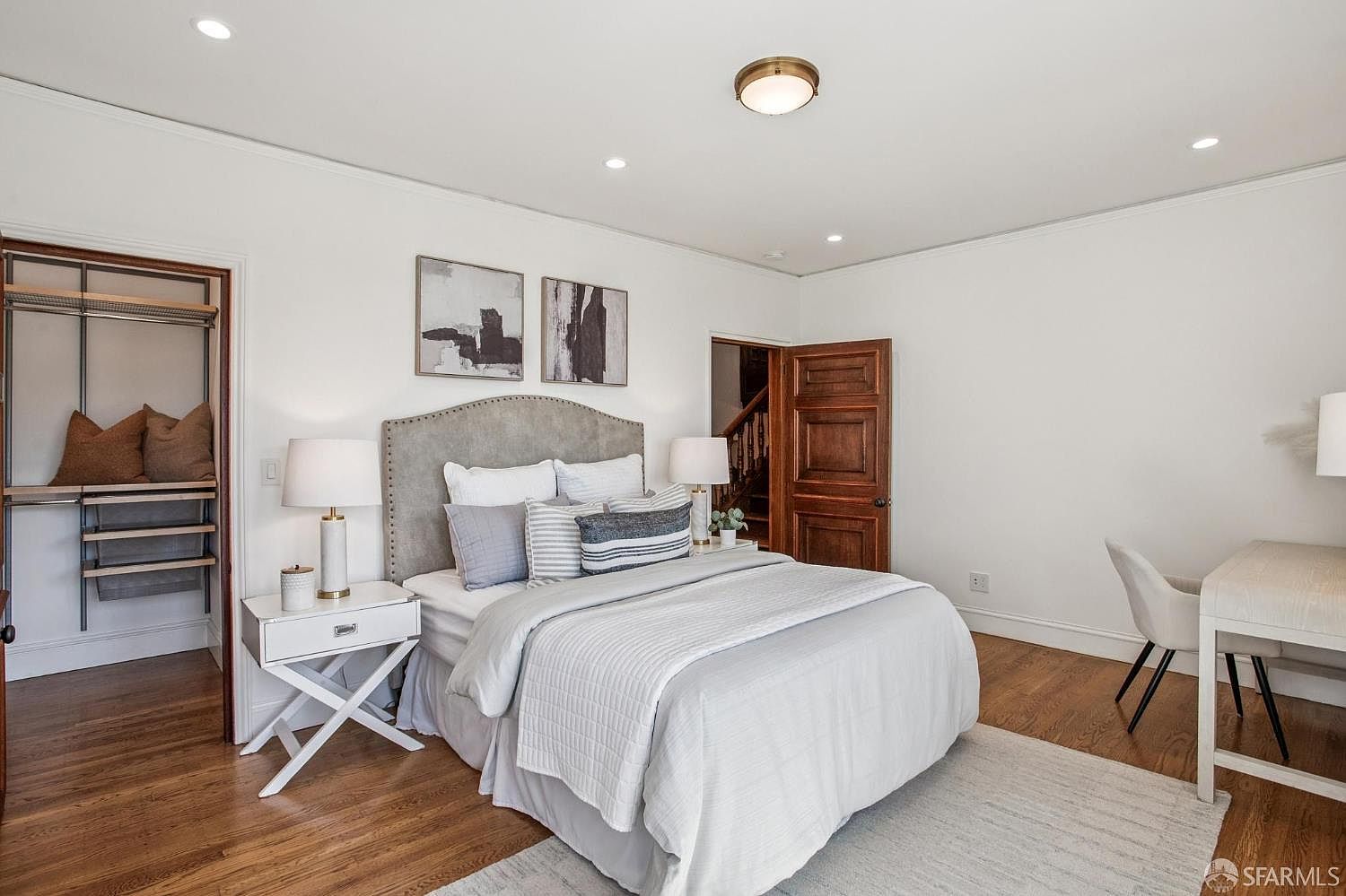This bright and airy primary bedroom features a neutral color palette with a plush upholstered headboard and layered bedding. To the left, an open-concept walk-in closet provides convenient storage, while a dedicated desk area on the right offers a functional workspace. The room is finished with warm wood flooring and recessed lighting, creating a clean and inviting atmosphere.