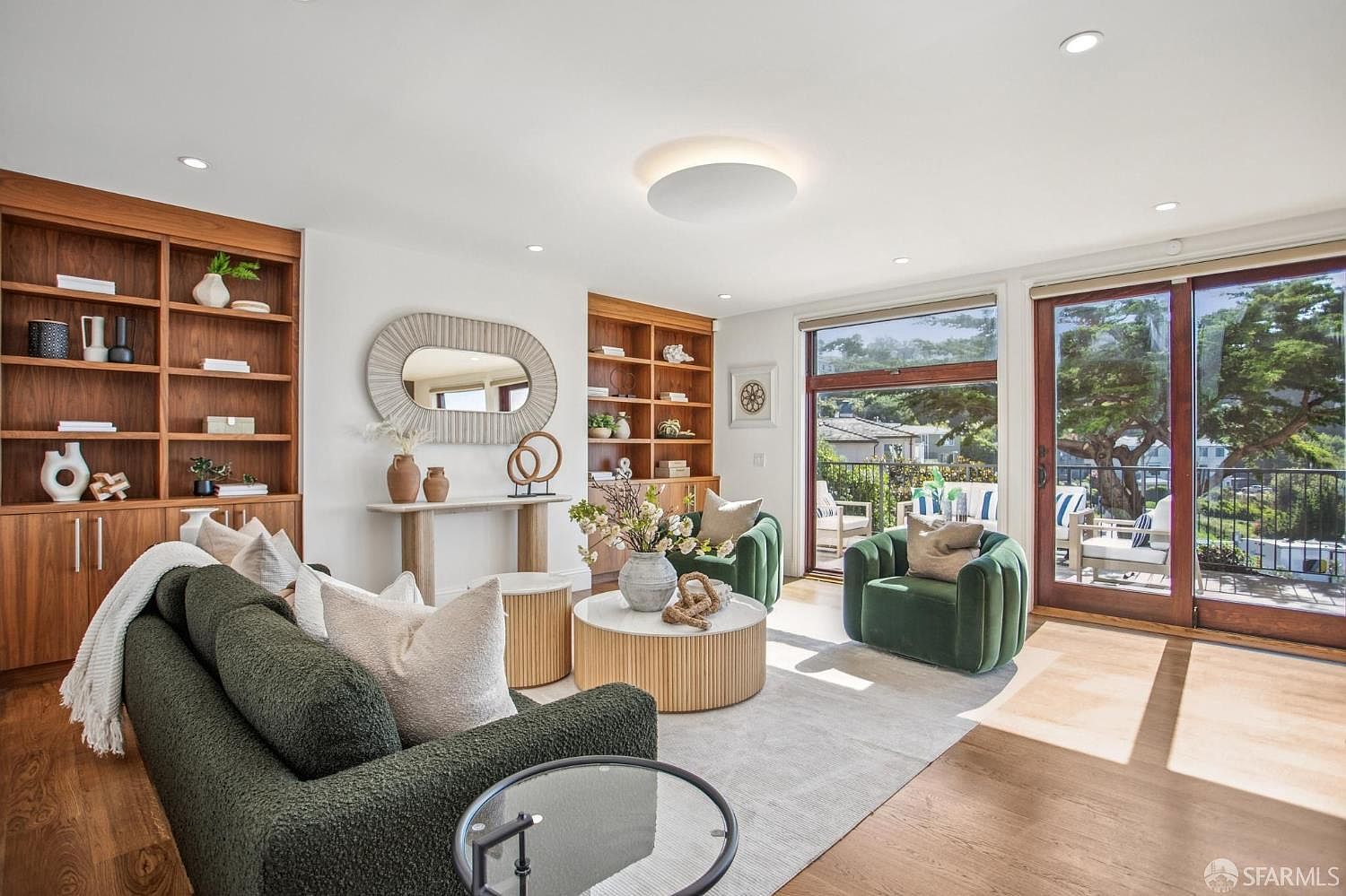 This bright and airy living room features a sophisticated, neutral color palette with textured green accent furniture. The space is anchored by built-in light wood shelving units on either side of a console table, while large glass sliding doors provide seamless access to an outdoor balcony. The room is bathed in natural light, highlighting the warm wood flooring and the contemporary circular ceiling light fixture.
