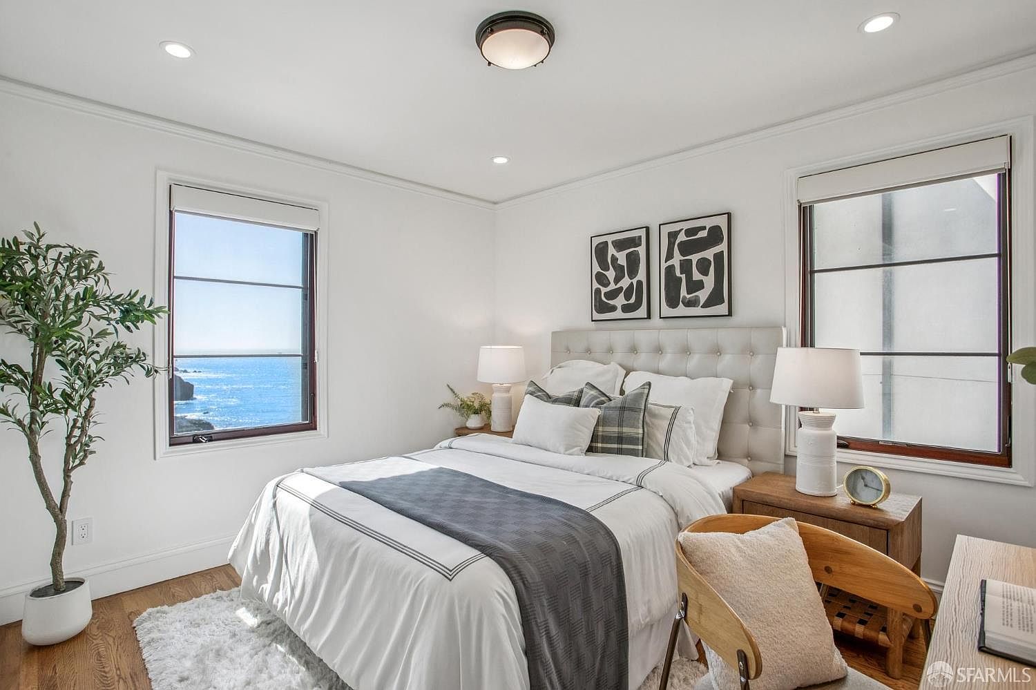 This bright and airy bedroom features a neutral color palette with a tufted headboard and layered bedding. Large windows on either side of the bed provide ample natural light and a stunning, unobstructed view of the ocean. The room is accented by a potted plant, a wooden chair, and minimalist wall art, creating a serene and inviting atmosphere.