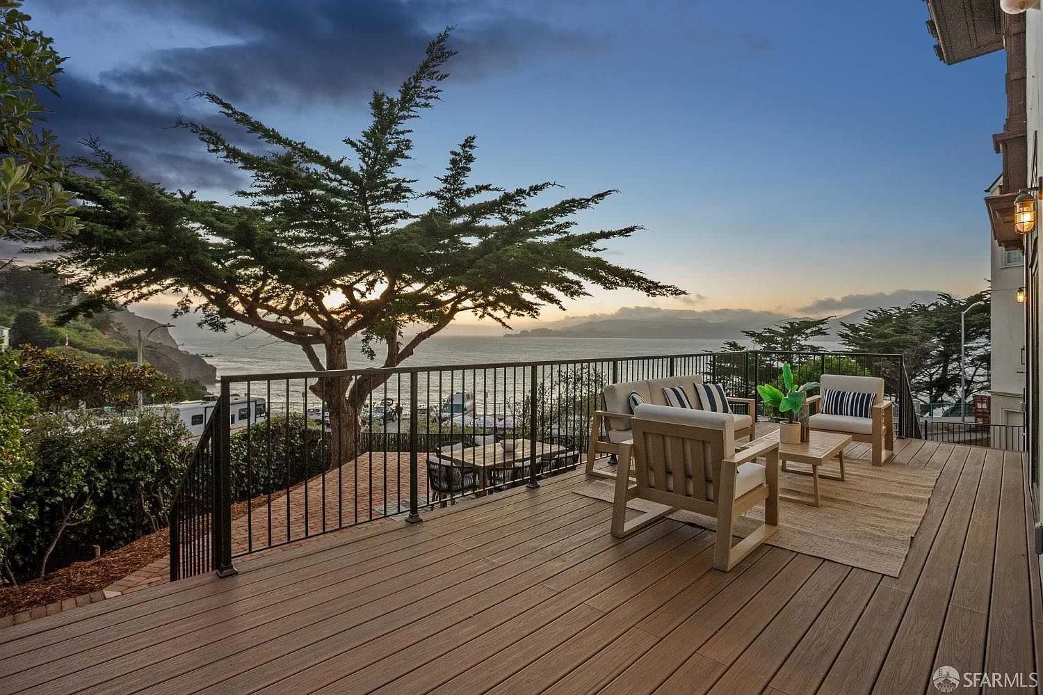 This expansive wooden deck offers a serene outdoor living space with a panoramic view of the ocean and a large, mature tree. The area is furnished with comfortable lounge seating, including a sofa and armchairs arranged on a textured rug, perfect for enjoying the sunset. A black metal railing provides safety while maintaining an unobstructed view of the coastal landscape below.