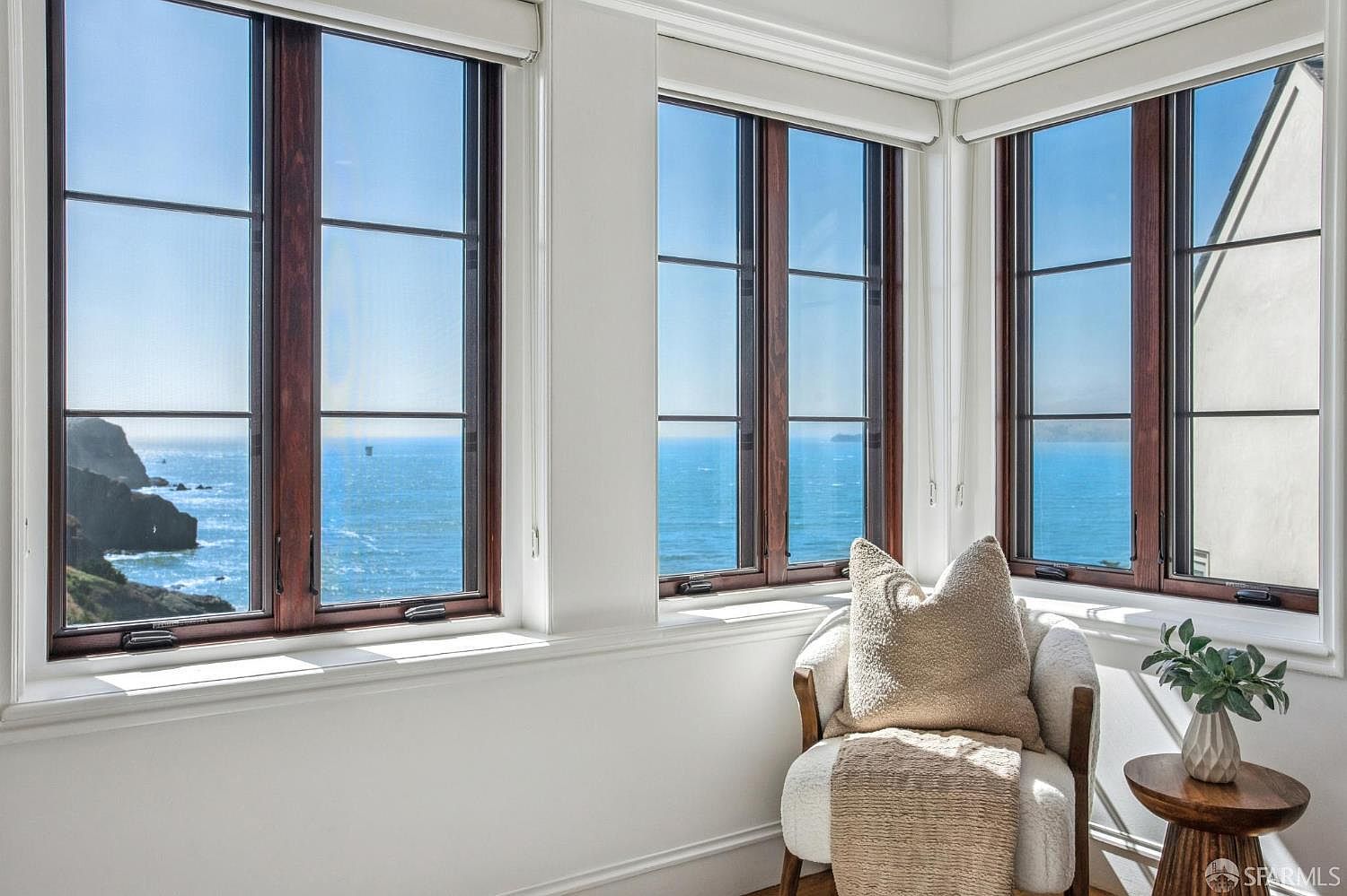 This bright corner nook features large windows with dark frames that offer expansive, panoramic views of the ocean and coastline. A cozy, textured armchair with a plush pillow and a draped throw sits beside a small, round wooden side table topped with a potted plant. The space is defined by crisp white walls and trim, creating a serene and inviting atmosphere perfect for relaxation.