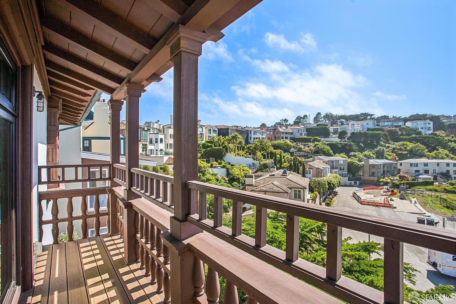 This image captures a wooden balcony with a classic balustrade railing, offering an elevated perspective of a suburban neighborhood. The structure features dark-stained wood posts and beams, creating a warm, traditional aesthetic that frames the expansive view of rolling hills and residential homes. The scene is bathed in bright, natural daylight, highlighting the clear blue sky and the surrounding greenery.