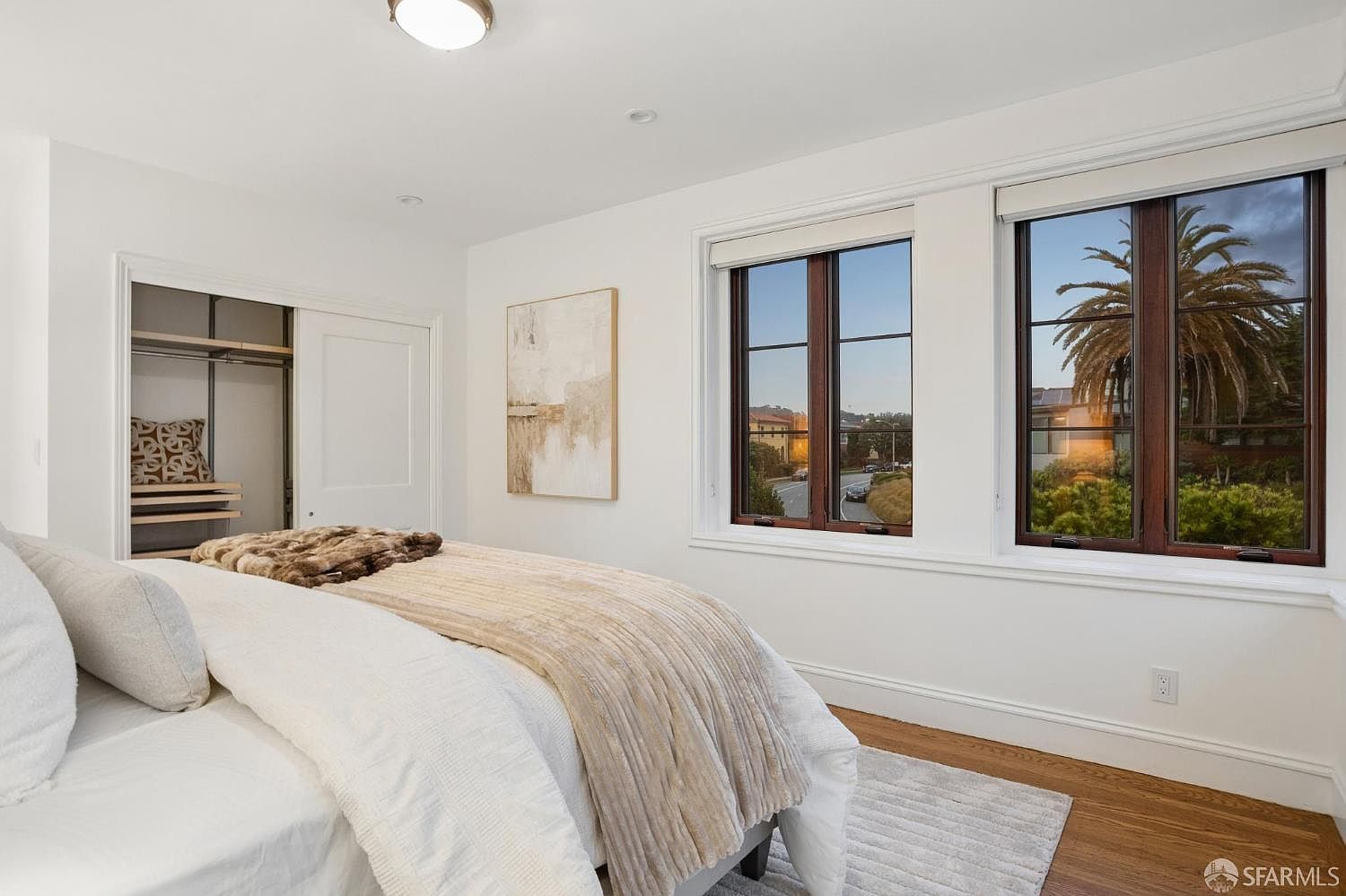 This bright and airy bedroom features crisp white walls, light wood flooring, and a plush bed dressed in neutral-toned linens. Two large windows with dark wood frames provide ample natural light and a view of the surrounding neighborhood, while a sliding-door closet offers convenient storage. A piece of abstract wall art adds a subtle decorative touch to the minimalist, clean aesthetic of the room.