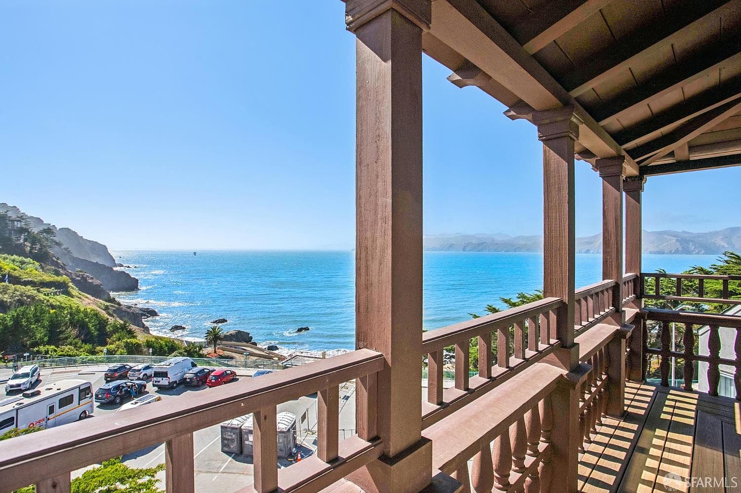 This image captures a scenic view from a wooden balcony overlooking a vast, blue ocean and rugged coastline. The balcony features sturdy wooden railings and posts, providing a classic, rustic frame for the expansive coastal vista. Below, a parking area with several vehicles is visible, nestled between the hillside and the water, creating a striking contrast between the natural landscape and human activity.