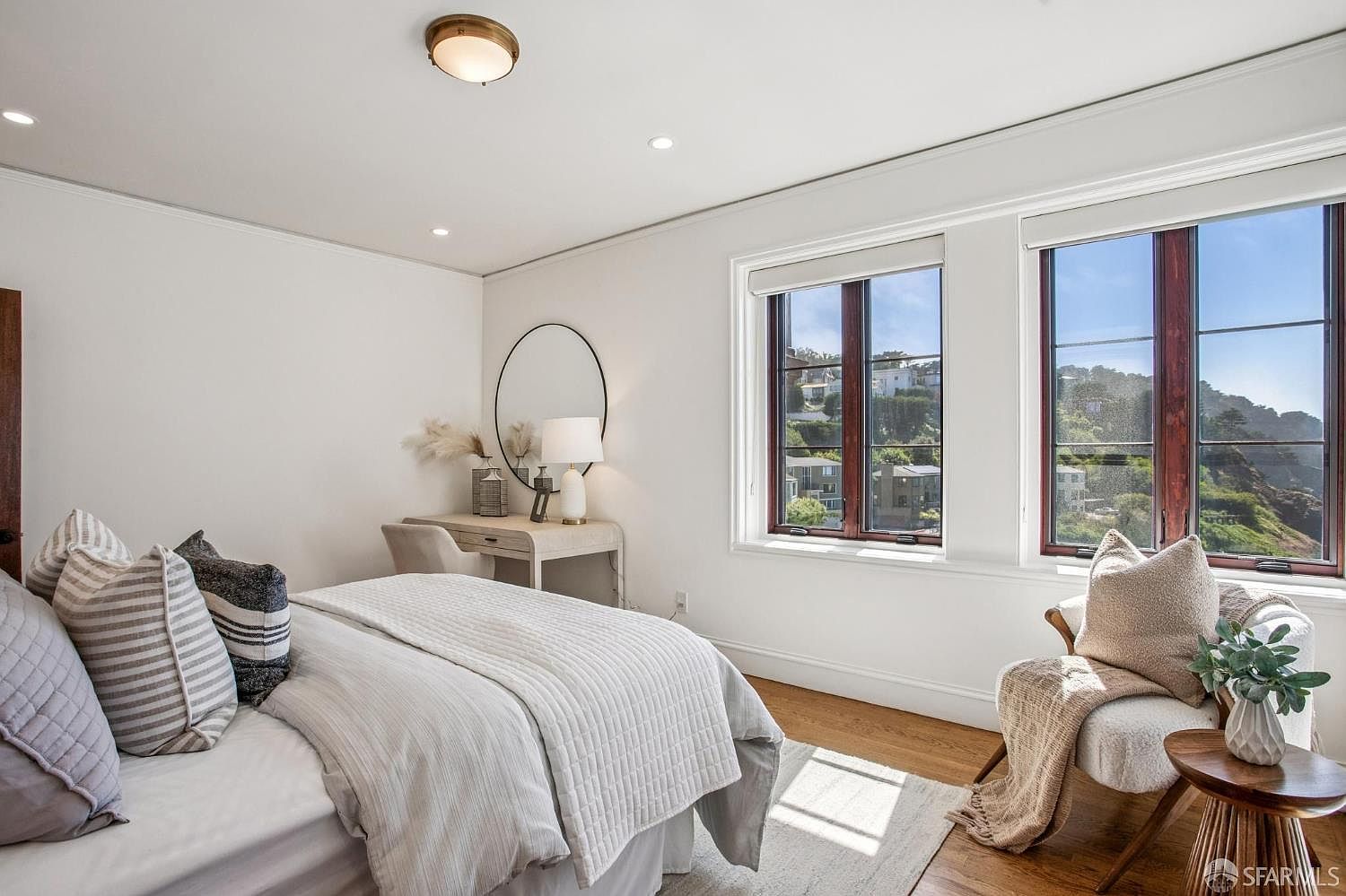 This bright and airy bedroom features a neutral color palette with soft textures and light wood flooring. A vanity area with a round mirror and lamp sits against the wall, while a cozy armchair with a throw blanket is positioned near large windows that offer a scenic view of the surrounding hills. The room is bathed in natural light, creating a serene and inviting atmosphere.