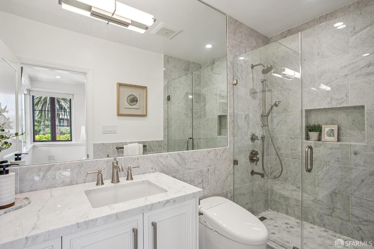 This bright and modern bathroom features a white vanity with a stone countertop and a large, wall-to-wall mirror. To the right, a glass-enclosed walk-in shower is finished with light-toned stone tile and includes a built-in recessed shelf. A sleek, white toilet with an integrated bidet seat sits adjacent to the shower, creating a clean and cohesive aesthetic.