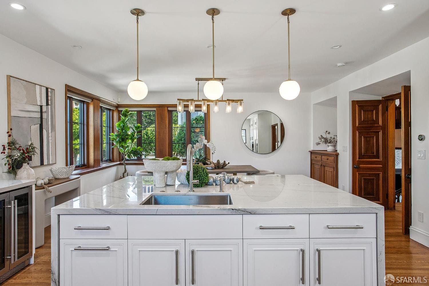 This bright and airy kitchen features a large central island with a stone countertop and an integrated sink, serving as the focal point of the room. The space is illuminated by three elegant pendant lights and recessed lighting, highlighting the white cabinetry and warm wood flooring. A dining area is visible in the background, complemented by large windows that bring in natural light and a circular wall mirror that adds depth to the open-concept design.