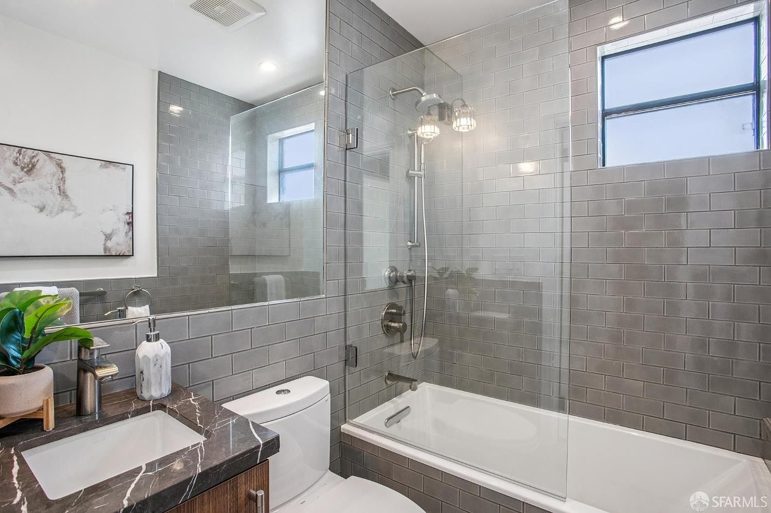 This modern bathroom features a sleek vanity with a dark stone countertop and a rectangular undermount sink. The shower-tub combination is enclosed by a glass panel and is finished with grey subway tiles that extend across the wall, creating a cohesive and contemporary aesthetic. A large mirror above the vanity reflects the space, while a small window provides natural light to the shower area.