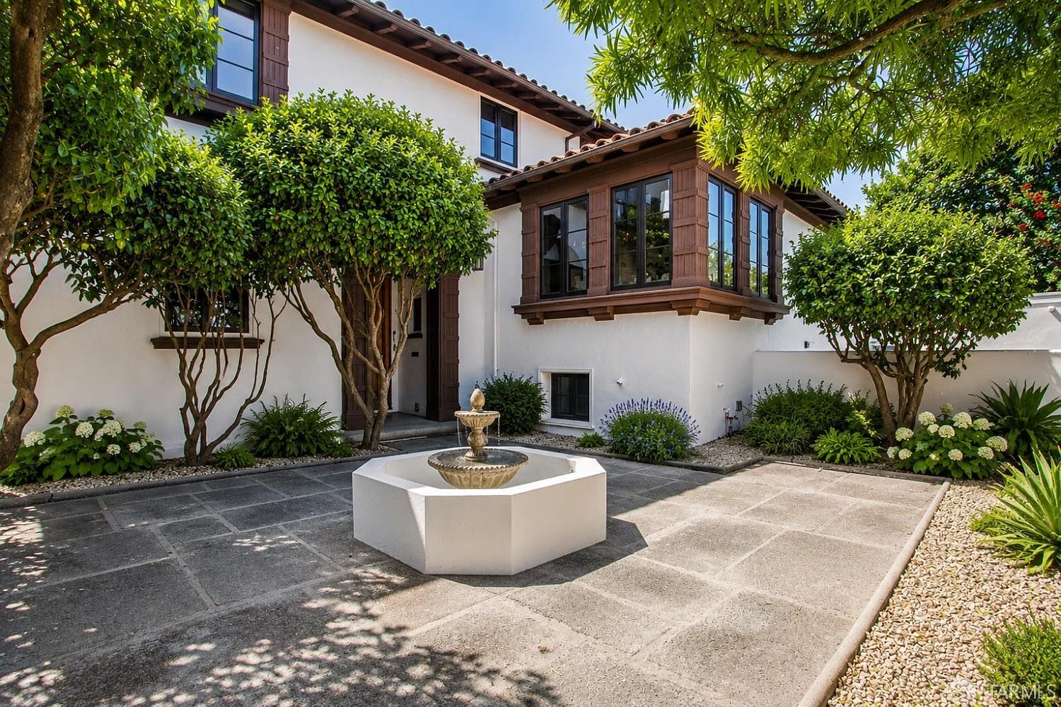 This charming courtyard features a central stone fountain set within a paved patio area, surrounded by lush greenery and mature trees. The home showcases a classic Mediterranean-inspired aesthetic with white stucco walls, dark wood-framed windows, and a tiled roof. The perspective captures a serene, private outdoor living space that seamlessly blends architectural elegance with natural landscaping.