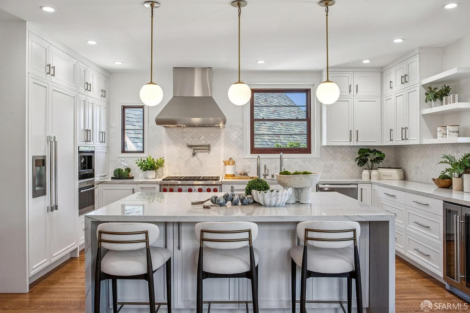 This bright and airy kitchen features a large central island with seating for three, topped with a light-toned stone surface. The space is equipped with white cabinetry, a stainless steel range hood, and a classic tile backsplash, all illuminated by three spherical pendant lights. The perspective is a centered, eye-level shot that highlights the clean, modern aesthetic and the functional layout of the workspace.